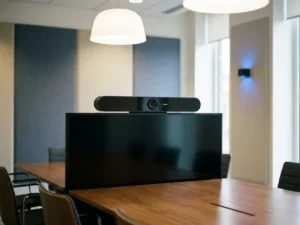 Hybride vergaderkamer camera op groot beeldscherm, met gepolijste conferentietafel en akoestische wandpanelen op de achtergrond.