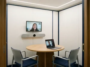 Compacte vergaderruimte met video-bar op klein scherm, ronde tafel met twee stoelen en laptop met remote deelnemer.