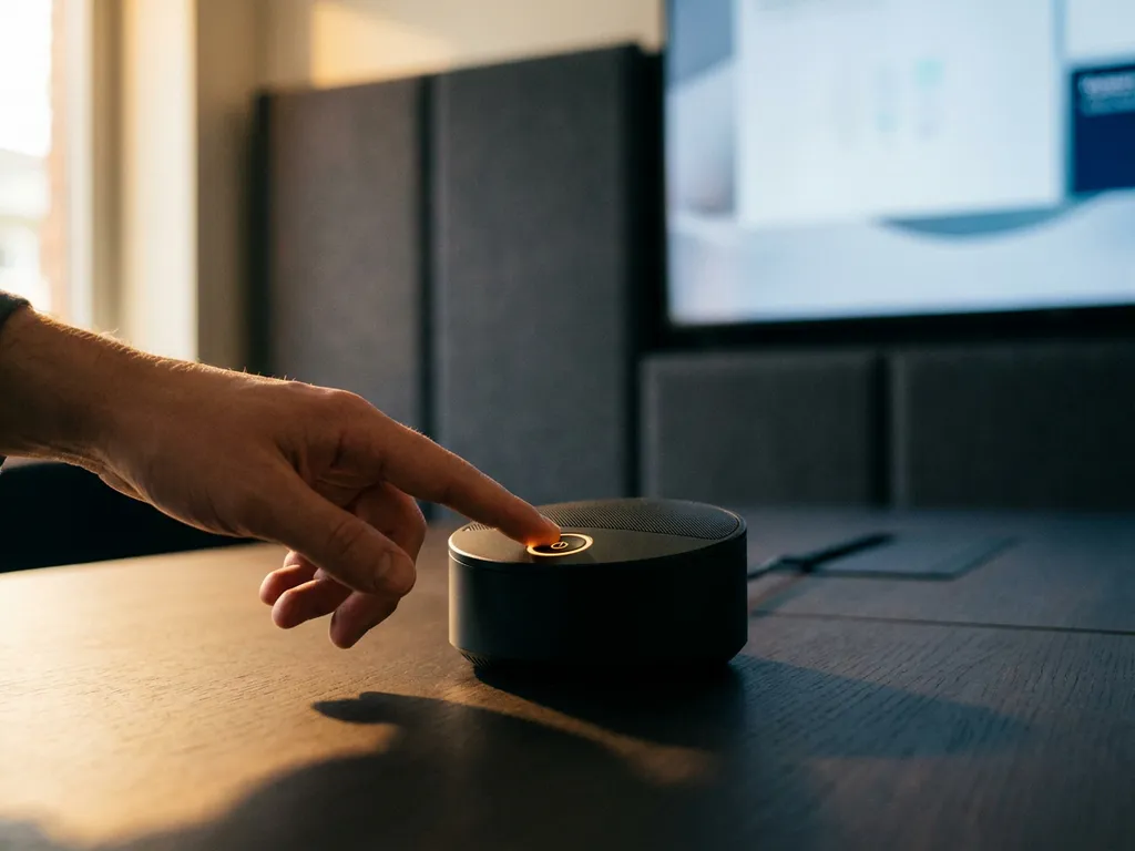 Hand die een verlichte standby-knop van een zwarte AI-microfoon op een vergadertafel aanraakt, met akoestische panelen op de achtergrond.