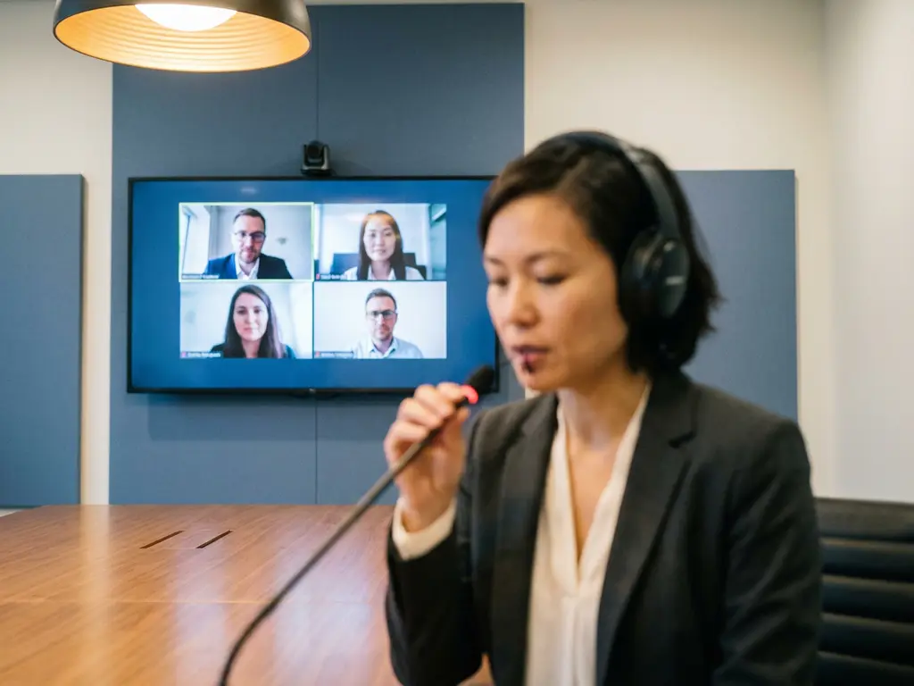 Professionele tolk met koptelefoon spreekt in microfoon tijdens hybride vergadering, groot scherm met deelnemers op afstand op achtergrond.